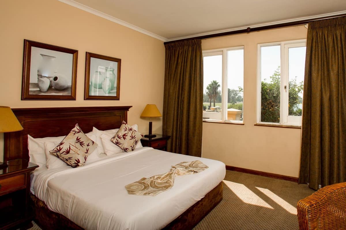 A hotel room with a large bed, two framed pictures on the wall, a nightstand with a lamp, and a view of the outdoors through the window.