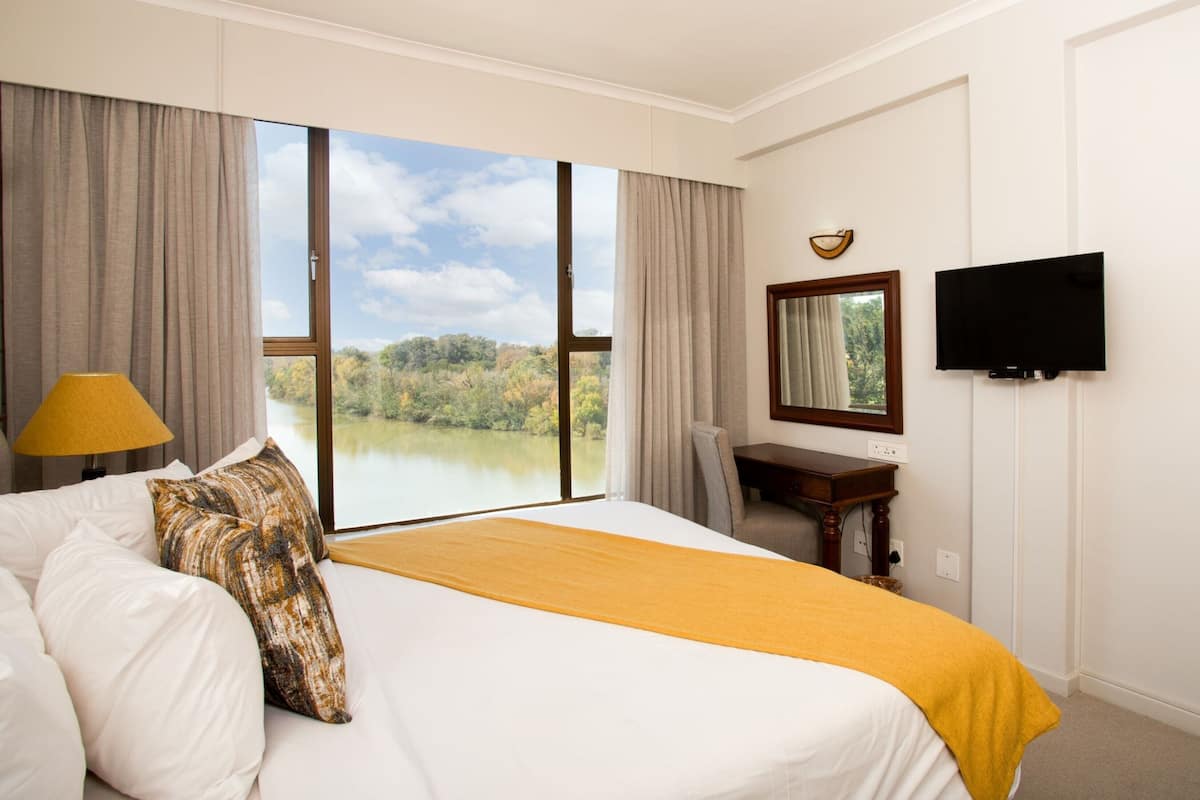 A hotel room with a large bed, a desk, a chair, a TV, and a view of a lake.