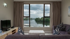 A living room with a large window overlooking a river, a flat-screen TV, a sofa, and a coffee table. - Riviera On Vaal Resort (Vereeniging)