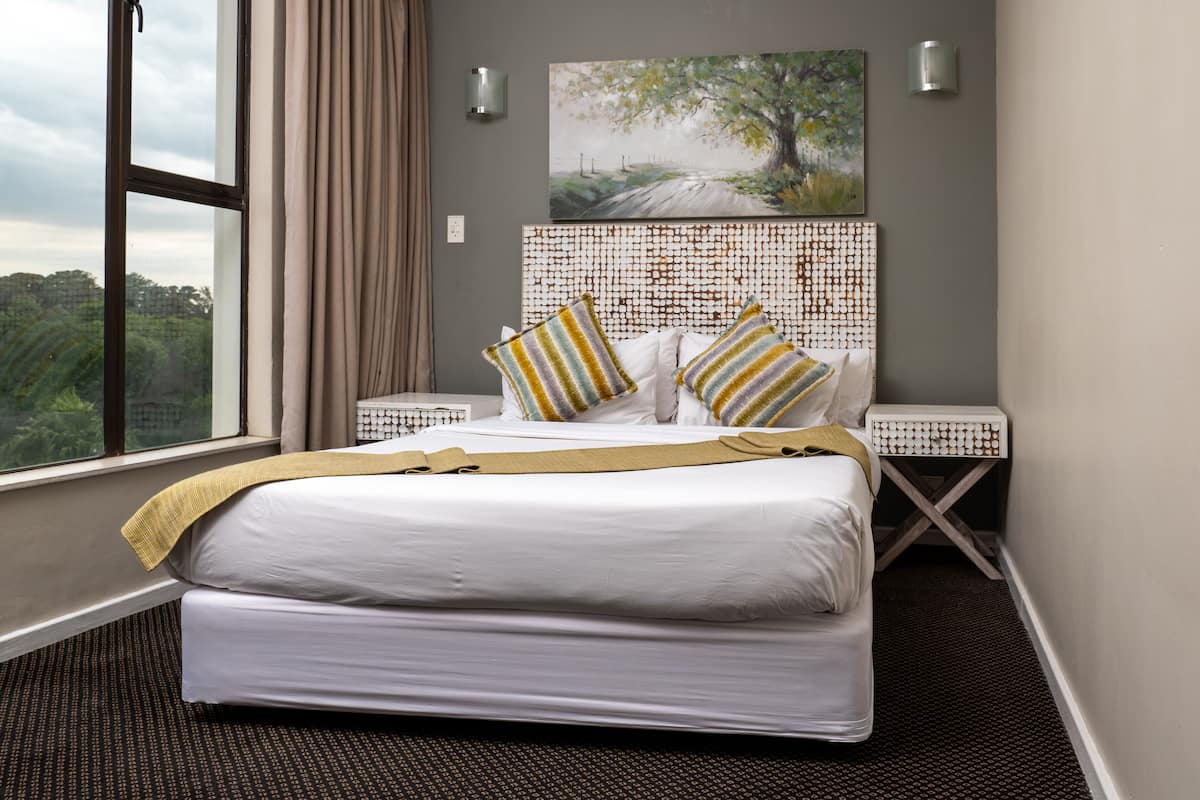A hotel room with a large bed, two bedside tables, a painting on the wall, and a view of greenery outside.
