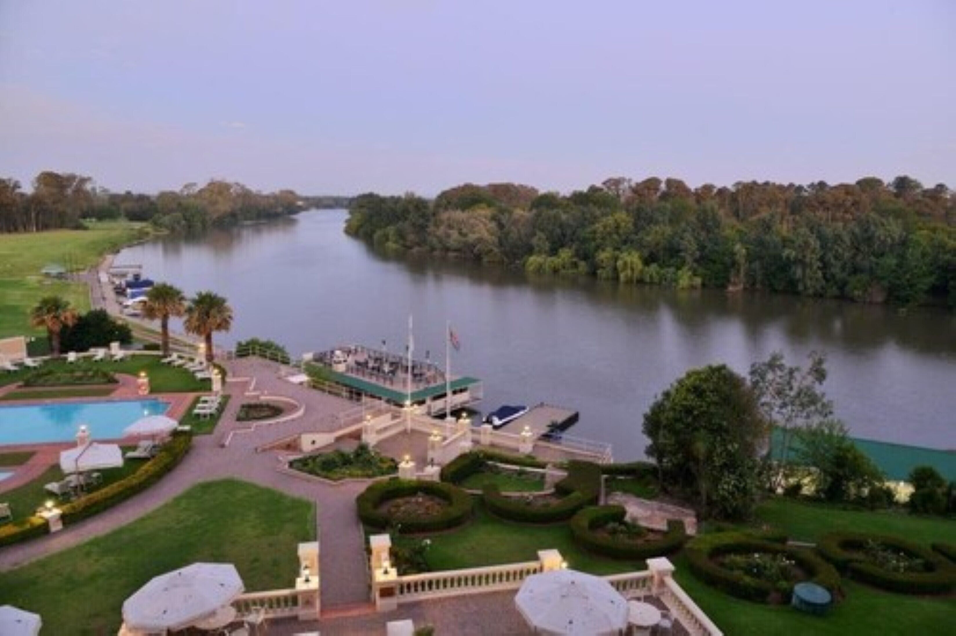 A riverside hotel with a swimming pool, landscaped gardens, and a central building with a flag.