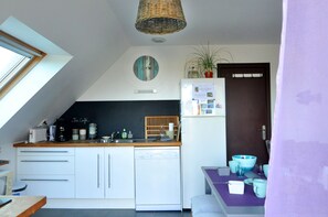 A compact kitchen with white cabinets, a small refrigerator, a wooden countertop, and a dining area with a purple table. - Cottage Maison Drogou (Porspoder)