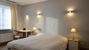 A bedroom with a large bed, a desk with a laptop, two bedside lamps, and a window with curtains.