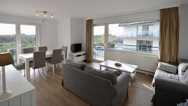 A modern living room with a grey sofa, a dining table with chairs, a TV, and large windows with curtains.