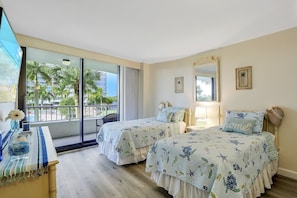 Quarto de hotel com duas camas, varanda e vista para palmeiras e edifícios.