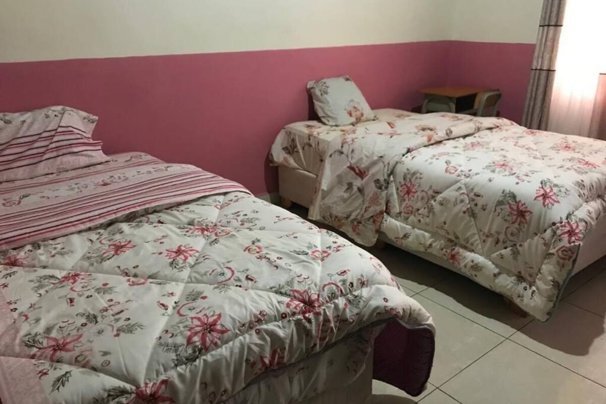 A bedroom with two beds, each with floral bedding, a pink wall, and a small table.