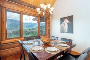 Un coin repas avec une table en bois dressée pour six personnes, de grandes fenêtres offrant une vue sur les montagnes et un lustre.