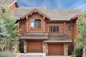 Une cabane en rondins avec des fondations en pierre, un grand garage à l’avant et une cheminée imposante.