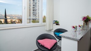 A modern room with a large window, a black chair with a pink cushion, a small table with a vase of flowers, and a view of a tall building and mountains. - Apartment Anica (Split)