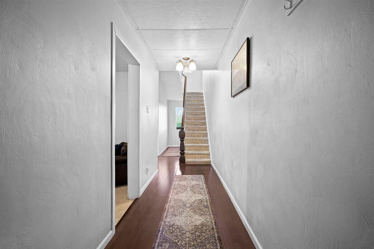 A hallway with a wooden floor, a patterned rug, a staircase, and a wall-mounted light fixture.