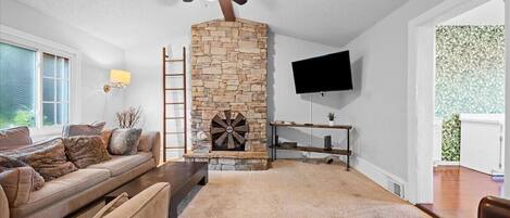 A living room with a stone fireplace, a ceiling fan, a TV mounted on the wall, and a sofa with cushions.