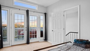 A bedroom with a bed, a window, a door, and a view of the outside.
