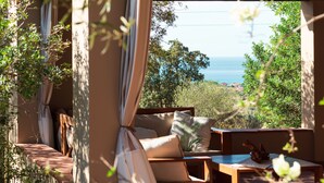 A sunlit outdoor seating area with a view of the sea, featuring a sofa, a table, and a chair.