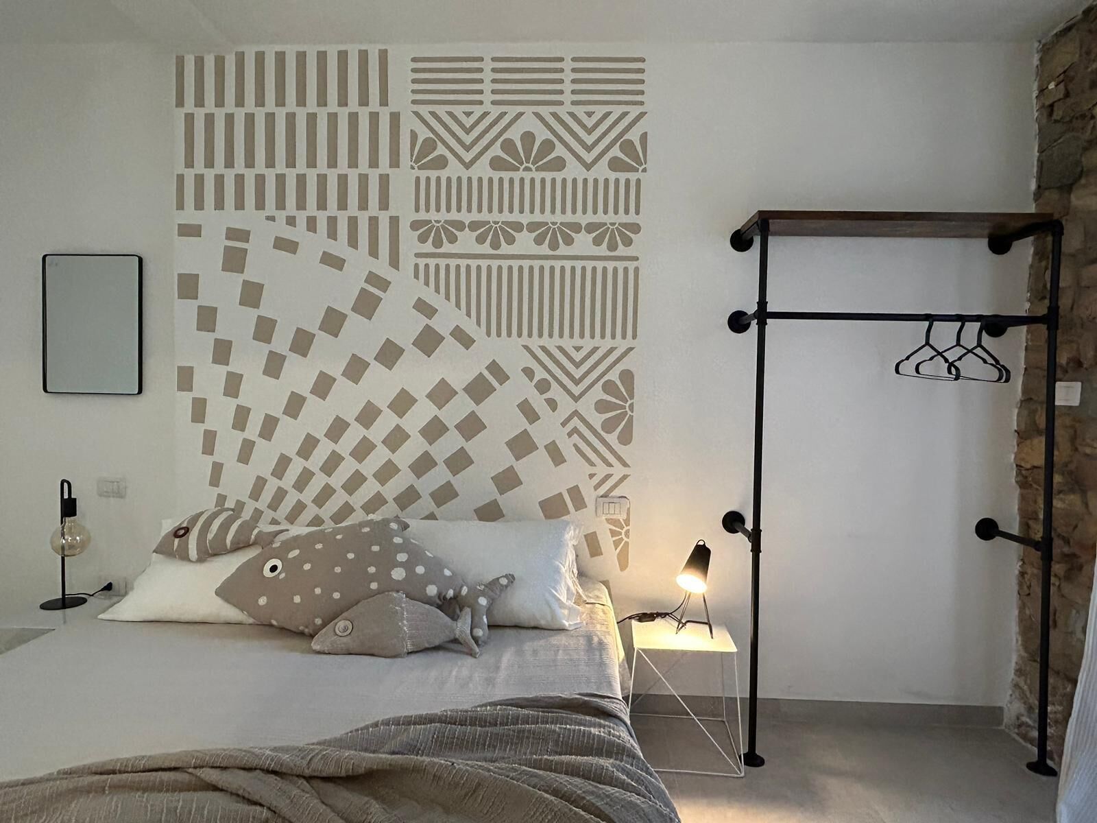 Een slaapkamer met een bed, kussens, een nachtkastje met een lamp, een muur met geometrische patronen en een kledingrek.