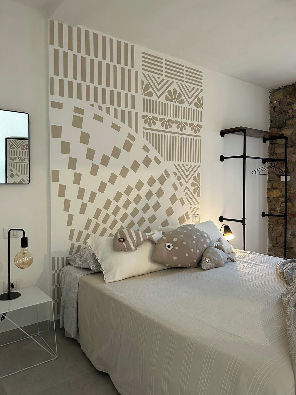 A bedroom with a patterned wall, a bed with pillows, a bedside table with a lamp, and a coat rack.