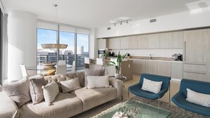 Un soggiorno moderno con un divano beige, poltrone blu e un tavolino in vetro. Si intravede una cucina con mobili in legno e un'area pranzo con un tavolo e sedie in vetro.