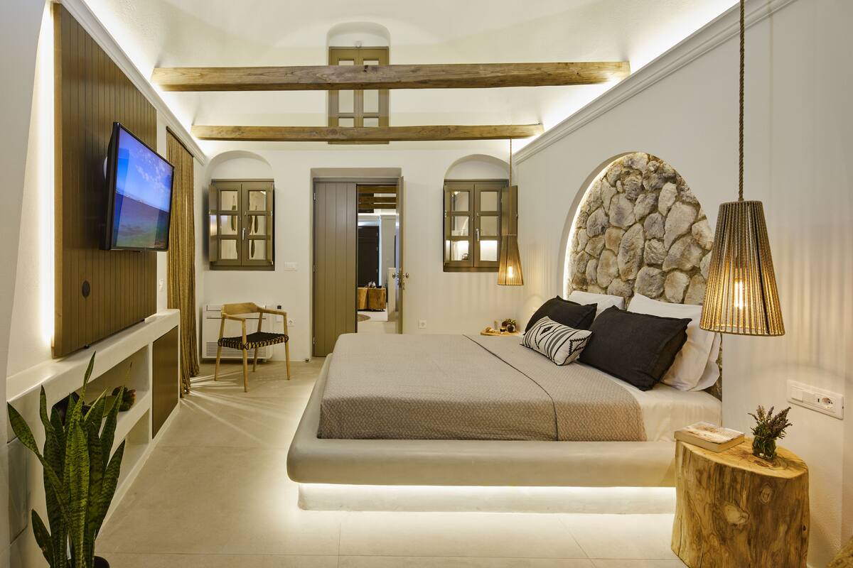 A bedroom with a bed, a TV mounted on the wall, a stone wall, and a wooden ceiling beam.