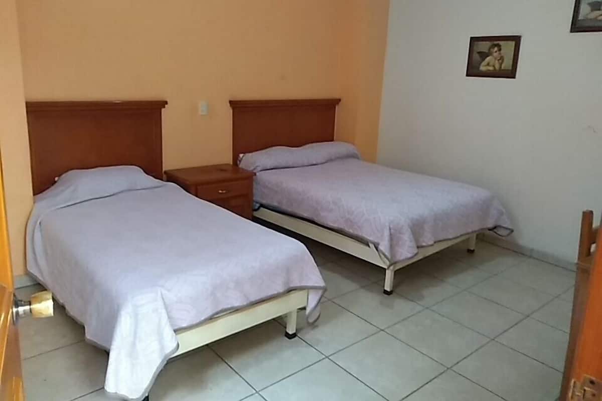 A small, clean room with two single beds, a wooden nightstand, and a tiled floor.