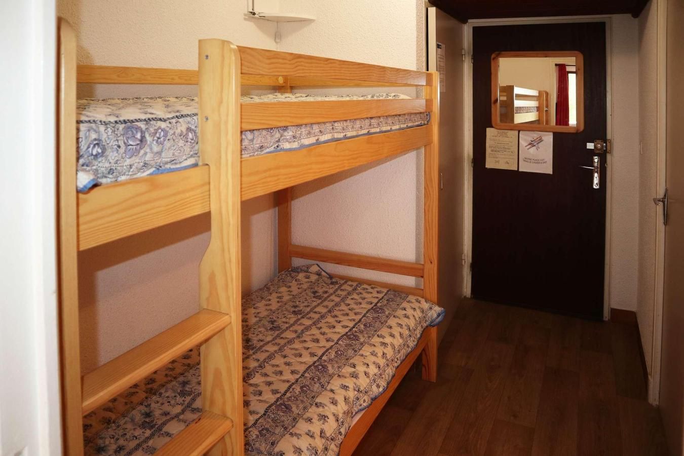 A bunk bed with patterned bedding, a door with a mirror, and a hallway with wooden flooring.