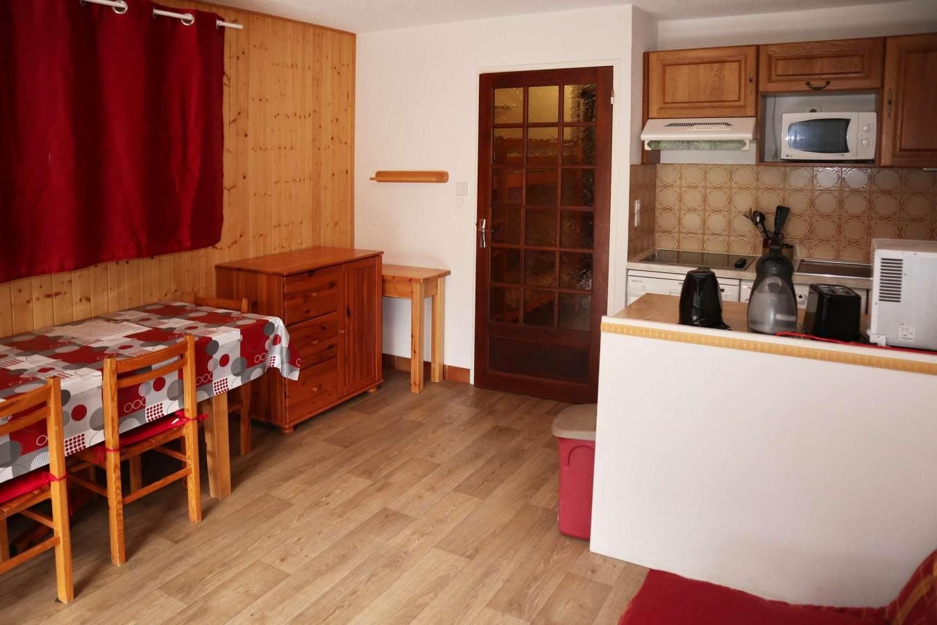 Une cuisine compacte avec des meubles en bois, un four à micro-ondes, une bouilloire et une petite table recouverte d’une nappe rouge.