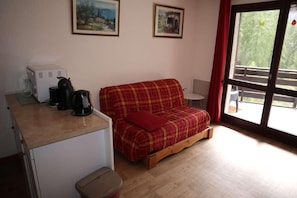 Ein kleiner Raum mit einer karoüberzogen Couch, einer Kaffeemaschine und einem kleinen Tisch. An der Wand hängen zwei gerahmte Bilder und ein Fenster mit Blick auf Grünflächen.