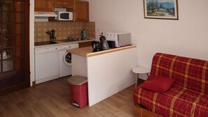 A compact kitchen with a microwave, sink, and washing machine. There is a small stool and a red trash bin. A red plaid sofa is in the living area. - 1st floor, balcony, 32m², Les Orres (Les Orres)