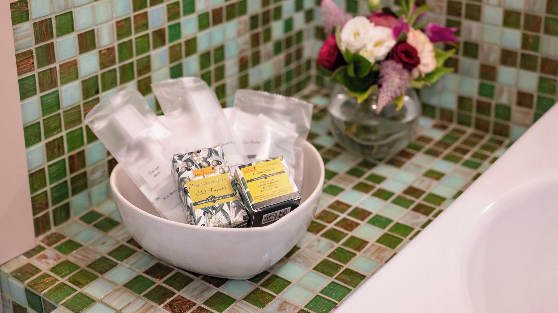 Un tazón blanco sobre una superficie de azulejos, con dos pastillas de jabón y un ramo de flores al fondo.