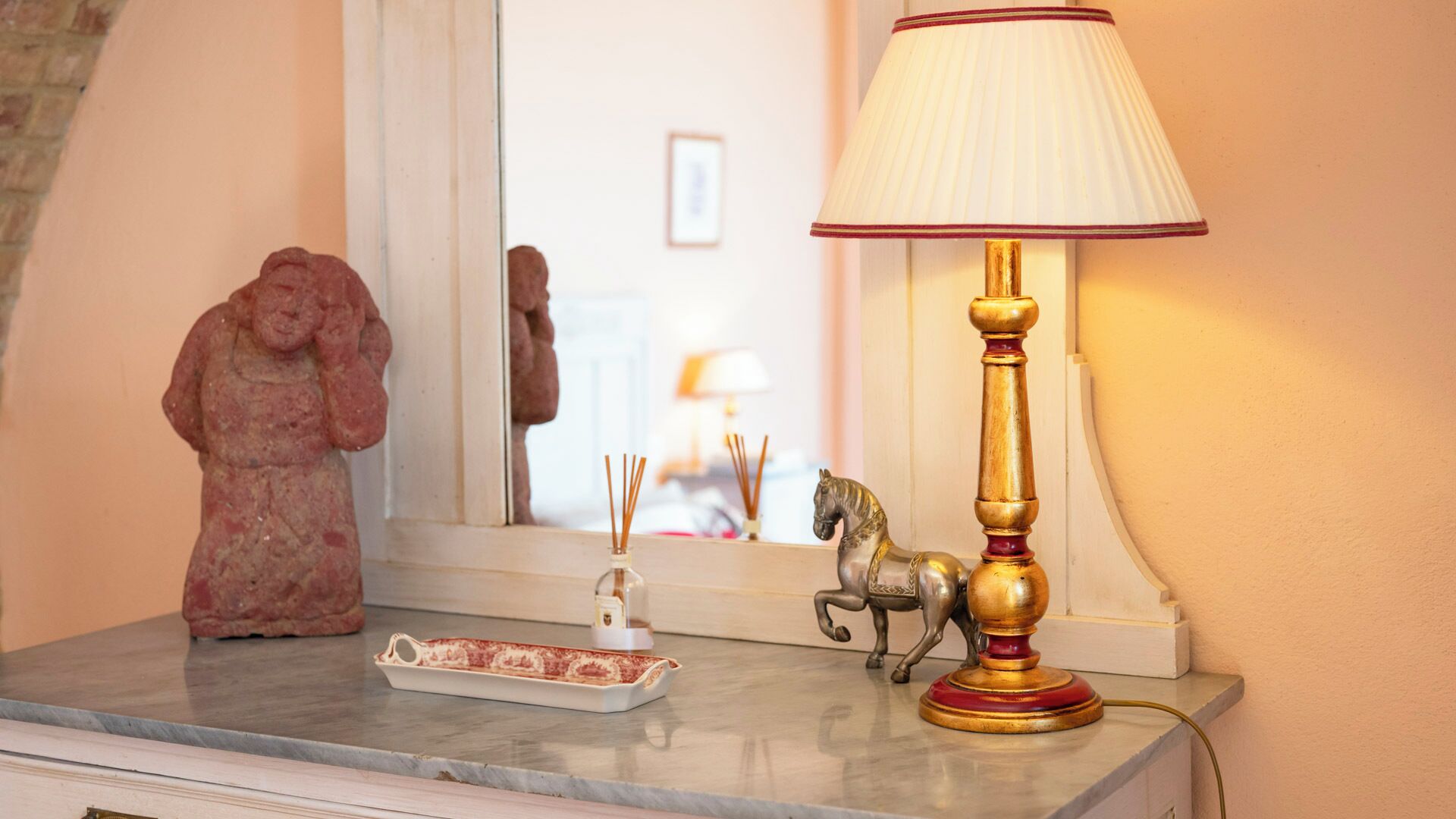Uma mesa com tampo de mármore, um abajur decorativo, incensos, uma pequena estatueta de cavalo e uma bandeja com um objeto vermelho.