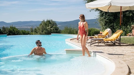 A swimming pool with clear blue water, a person standing on the edge, and another person swimming. There are lounge chairs and an umbrella in the background.