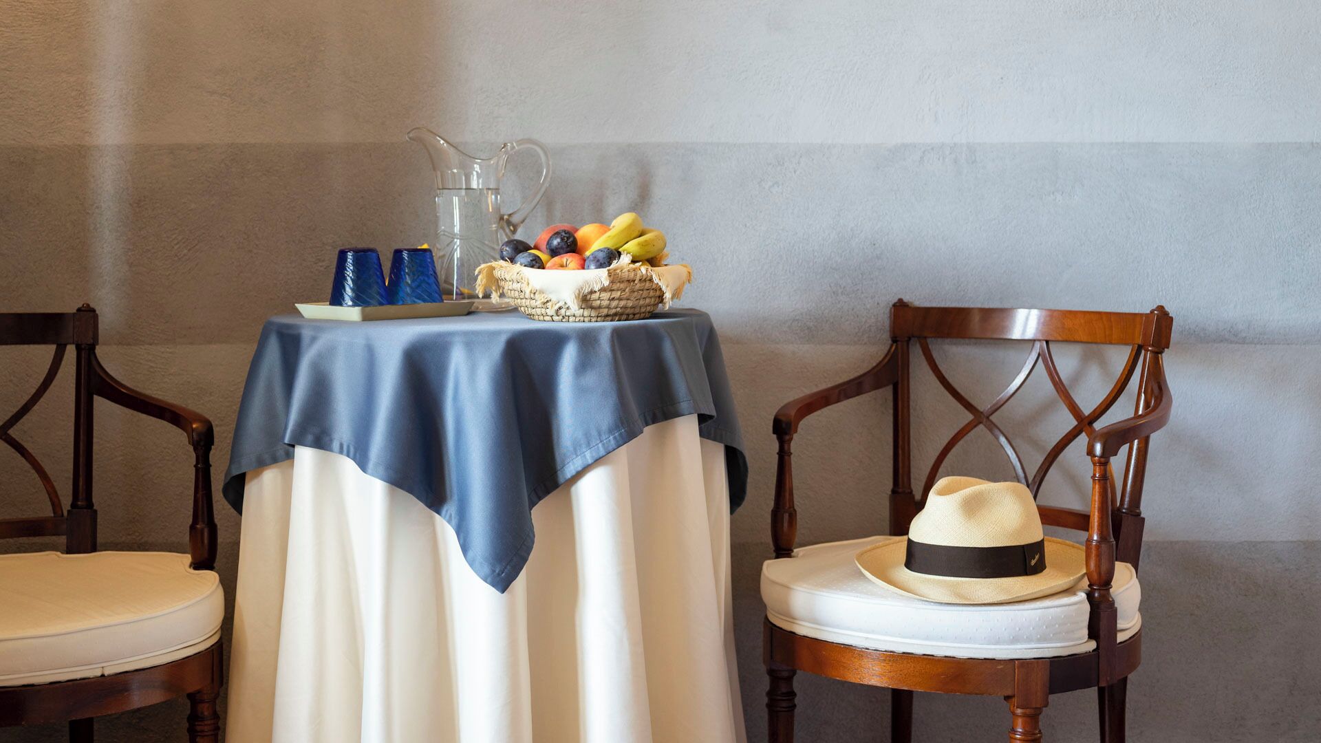 Una mesa con mantel azul, una jarrón y una cesta de frutas. Hay dos sillas de madera con cojines.