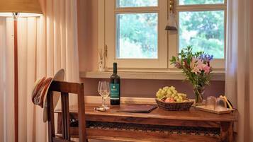 Une table en bois, avec une bouteille de vin, un verre, un panier de fruits et un vase de fleurs, près d’une fenêtre.