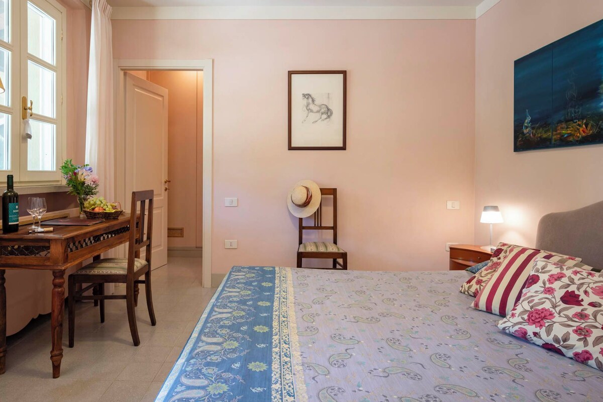 A bedroom with a bed, a desk with a chair, a lamp, a painting, and a door leading to another room.