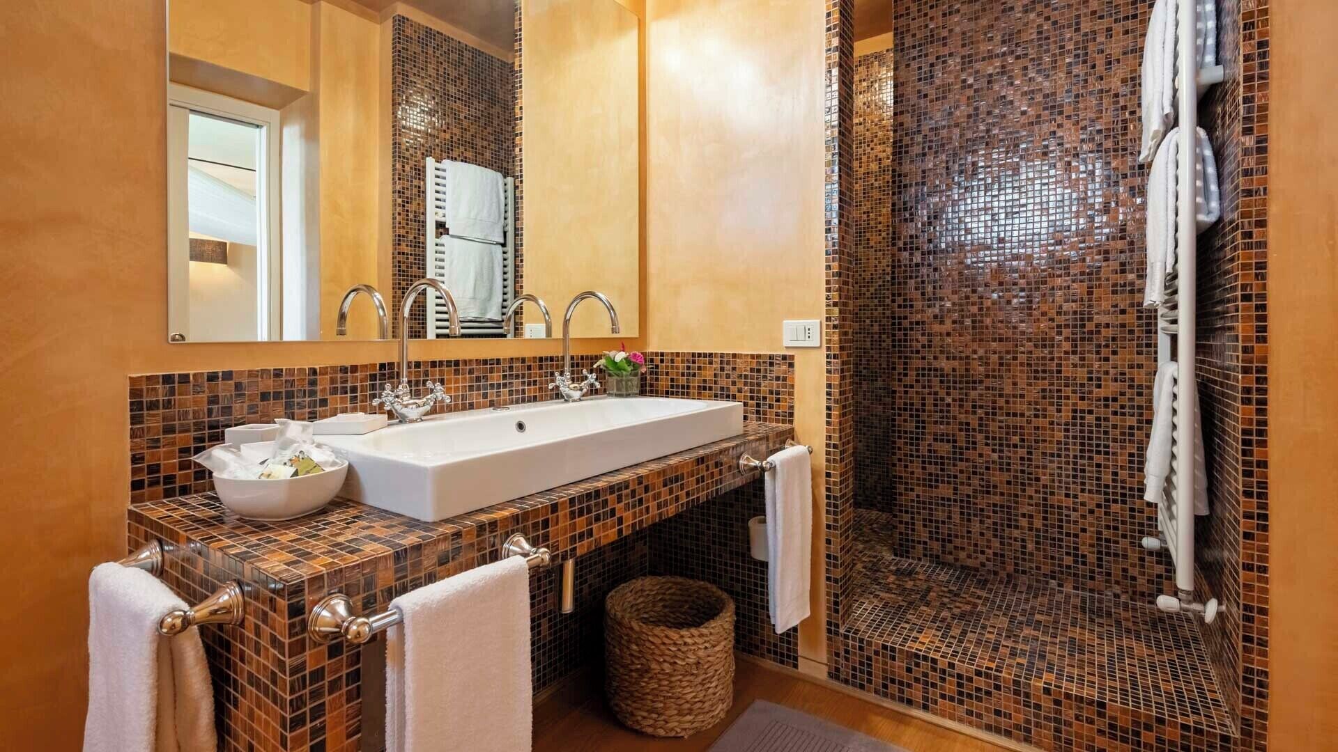 A bathroom with a large sink, two faucets, a mirror, and a shower with mosaic tiles.