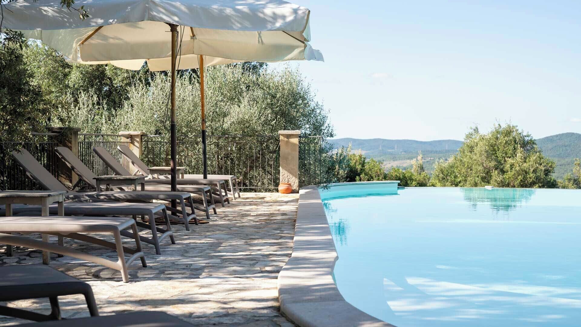 A poolside area with lounge chairs and a large umbrella, surrounded by stone flooring and greenery.