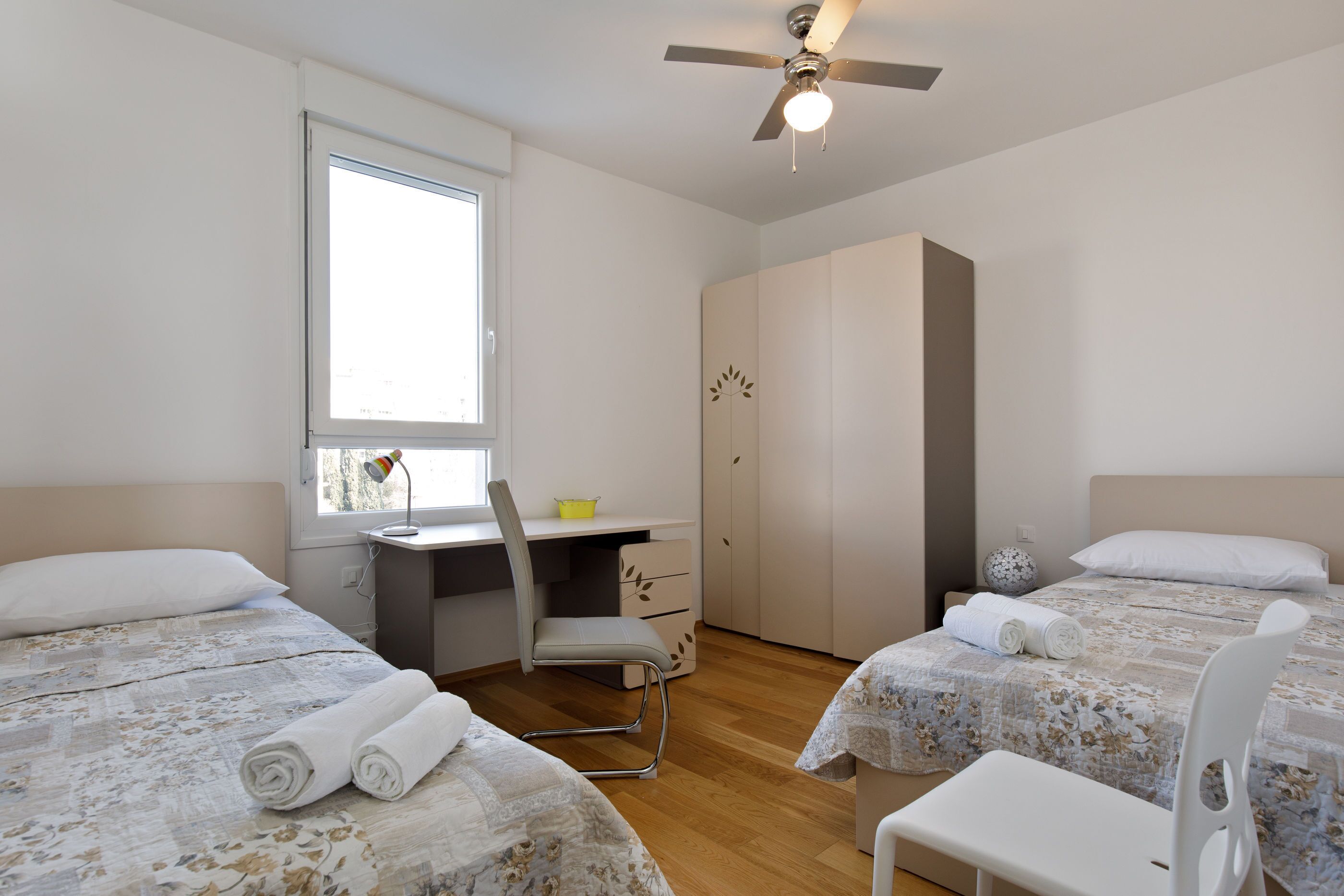 A bedroom with two beds, a desk, a chair, a wardrobe, and a ceiling fan.