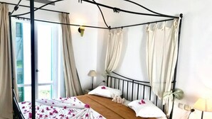 A four-poster bed with a canopy, white linens, a brown throw blanket, and pink flower petals on the bed. There is a nightstand with a lamp, a phone, and a small plant.