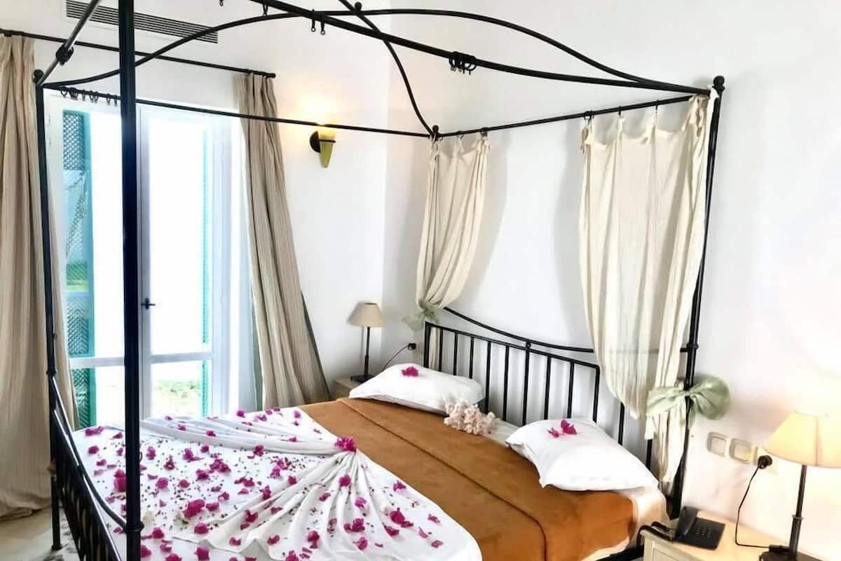 Una cama con dosel, ropa de cama blanca, una manta marrón y pétalos de flores rosadas sobre la cama. Hay una mesita de noche con una lámpara, un teléfono y una pequeña planta.
