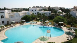 A large, curved swimming pool surrounded by lounge chairs and umbrellas, with white buildings and palm trees in the background.