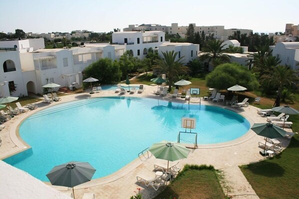 A large, curved swimming pool surrounded by lounge chairs and umbrellas, with white buildings and palm trees in the background.