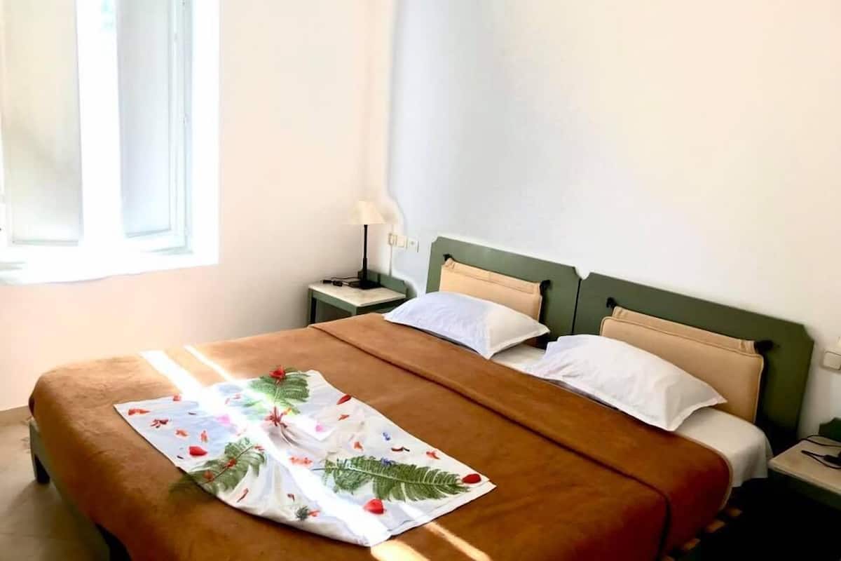 Una cama individual con una colcha marrón y un plaid estampado de flores. Hay una mesita de noche con una lámpara y una pequeña mesa junto a la cama. Se ve una ventana con una persiana blanca a la izquierda.
