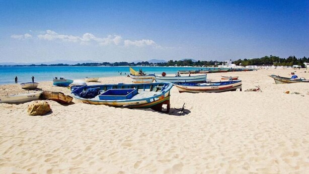 A beach with several small boats lined up on the sand, a clear blue sky, and a calm sea in the background.