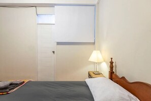 A bedroom with a bed, a nightstand with a lamp, a door, and a small window.