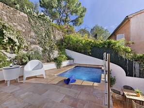 A small outdoor pool area with a stone wall, white chairs, a table, and a pergola.