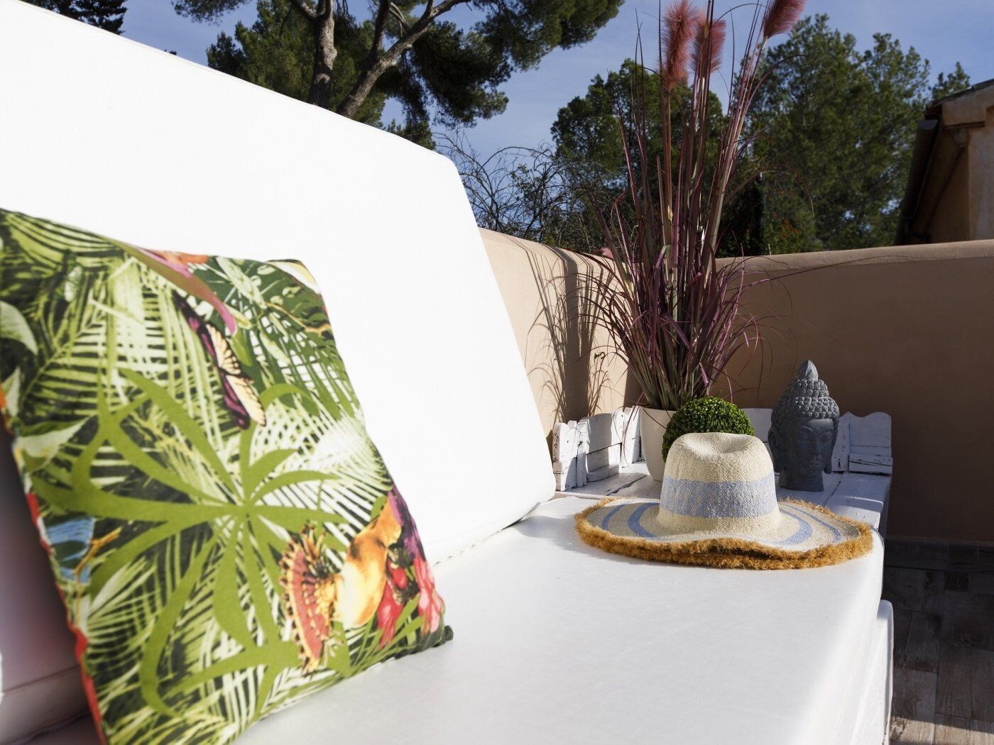 Un área de estar exterior blanca con un cojín de temática tropical, un sombrero de paja y plantas decorativas.