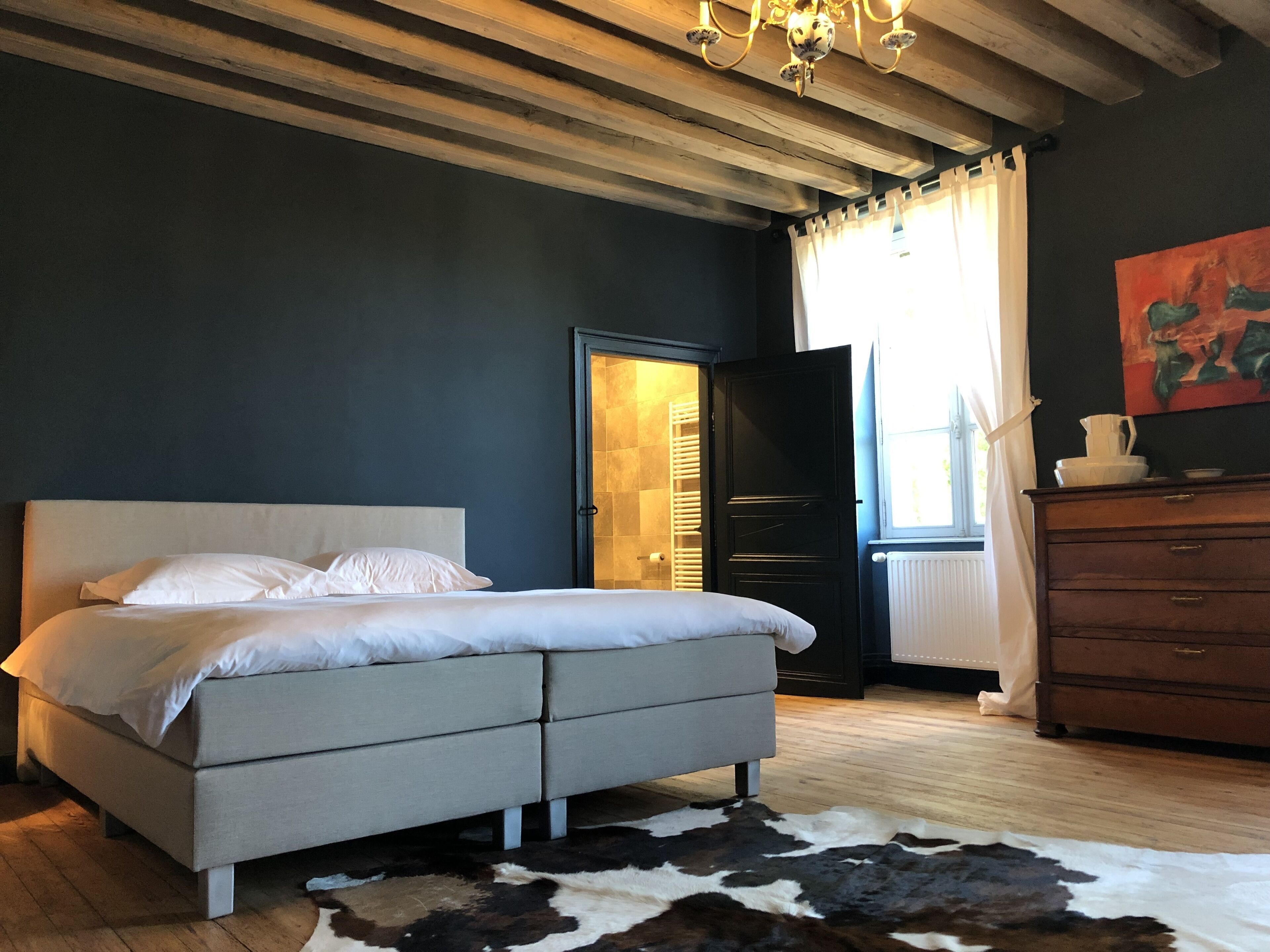 A bedroom with a large bed, a wooden ceiling, a chandelier, a cowhide rug, and a dresser.
