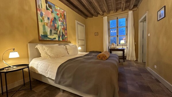 A bedroom with a large bed, a desk, a chair, a painting, and a window with curtains. - Zone Bleue (Hyds)