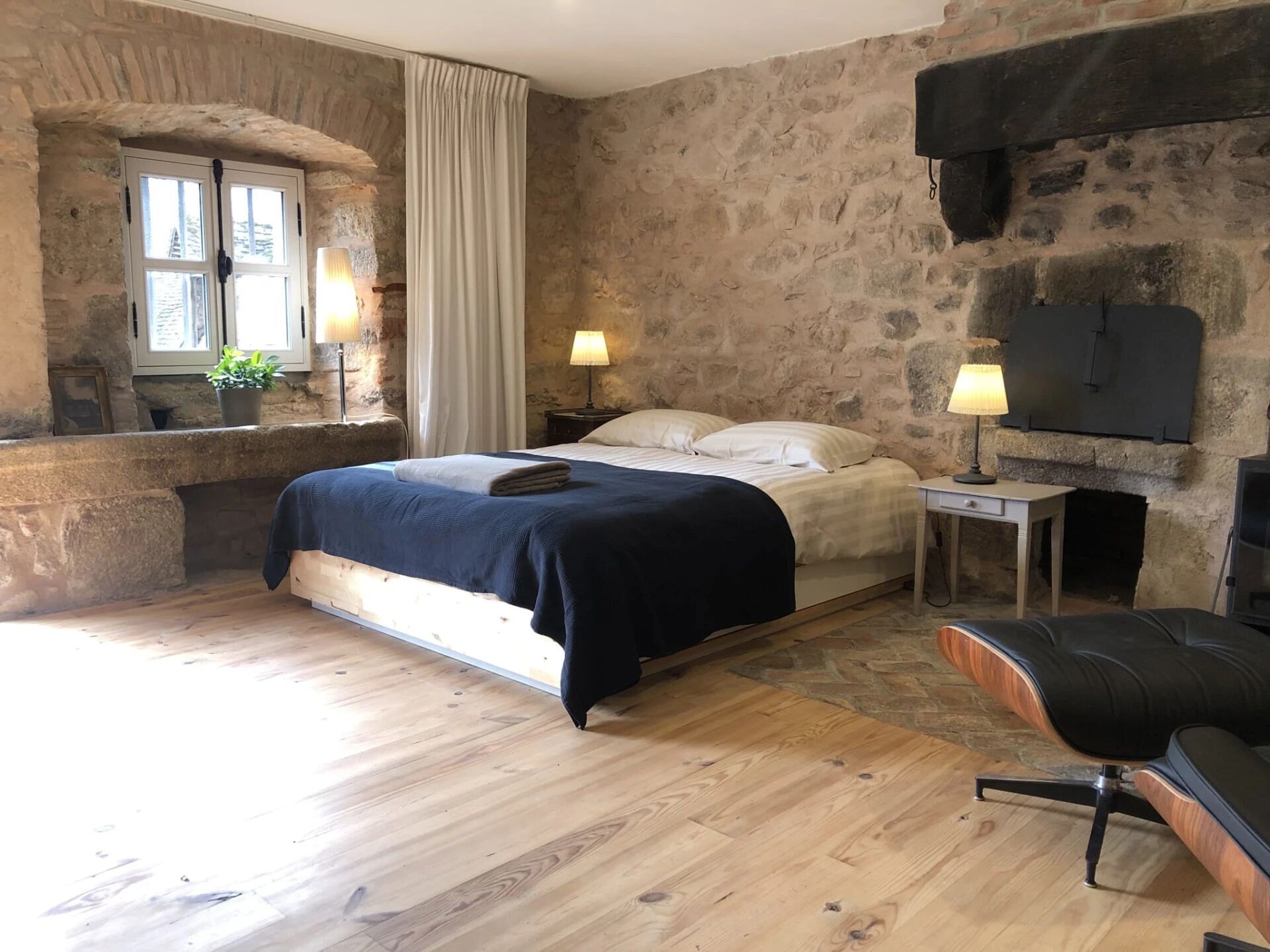 Um quarto com parede de pedra, cama com manta azul, soalho de madeira, uma cadeira, uma mesinha e uma televisão fixada na parede.