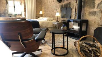 Une pièce chaleureuse avec une chaise en bois, un lit, une petite table, une lampe, une cheminée et un panier de bois de chauffage.