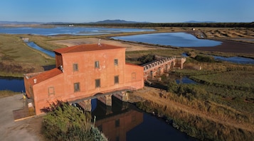 Un edificio grande de una sola planta con techo de tejas rojas, ubicado cerca de una masa de agua con varios estanques más pequeños y un puente que lo conecta a otra construcción.