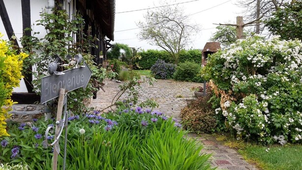 Un chemin de jardin menant à un bâtiment sur lequel est affiché un panneau « Bienvenue », le tout entouré d’une végétation luxuriante et de plantes en fleurs.